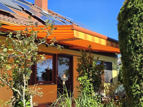 Haus mit orangefarbenem Sonnenschutz und Solarpanelen, umgeben von Bäumen und Pflanzen.